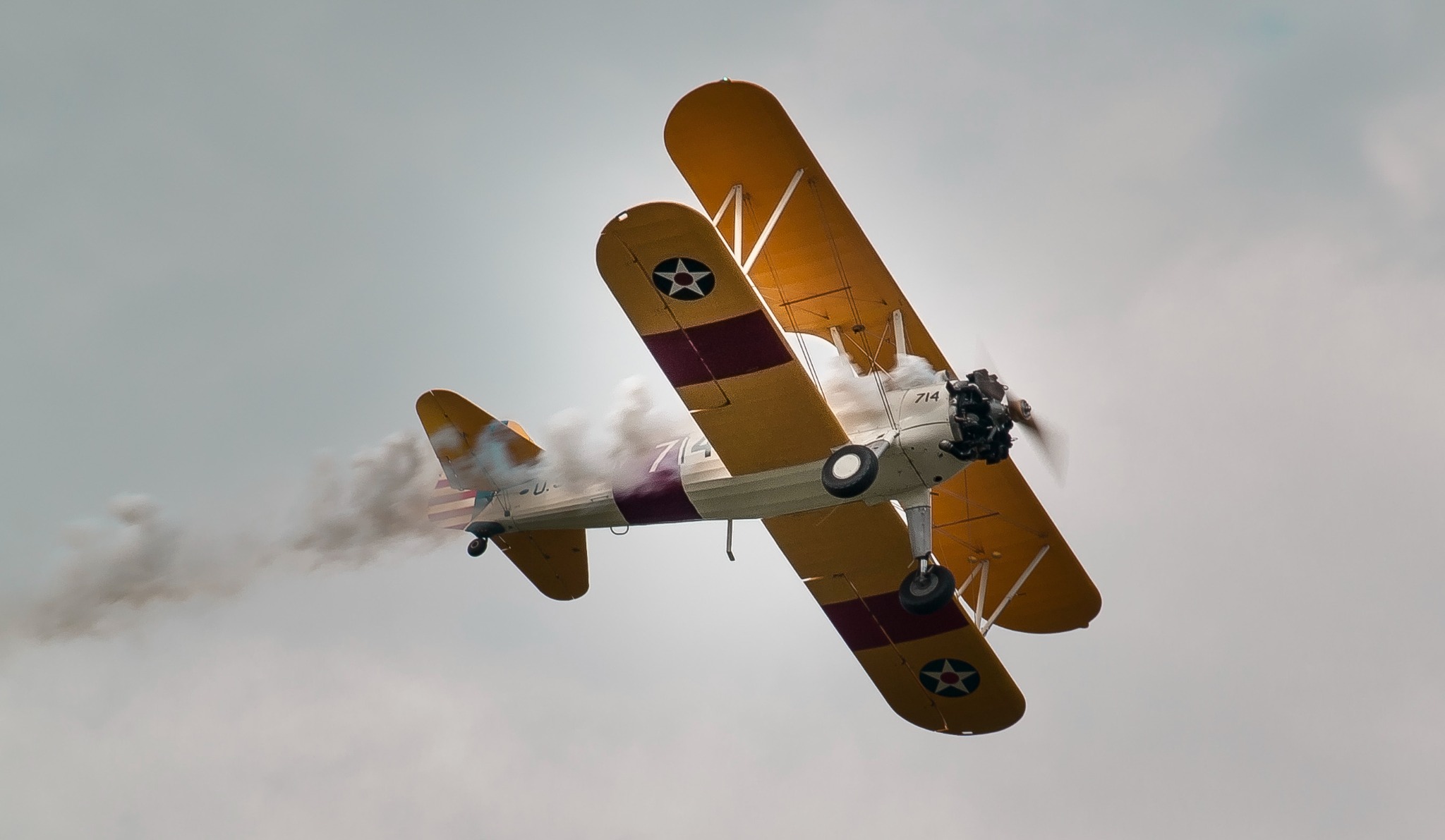 Żółty dwupłatowiec z numerem 714 i dymem wydobywającym się z silnika w locie na tle pochmurnego nieba.