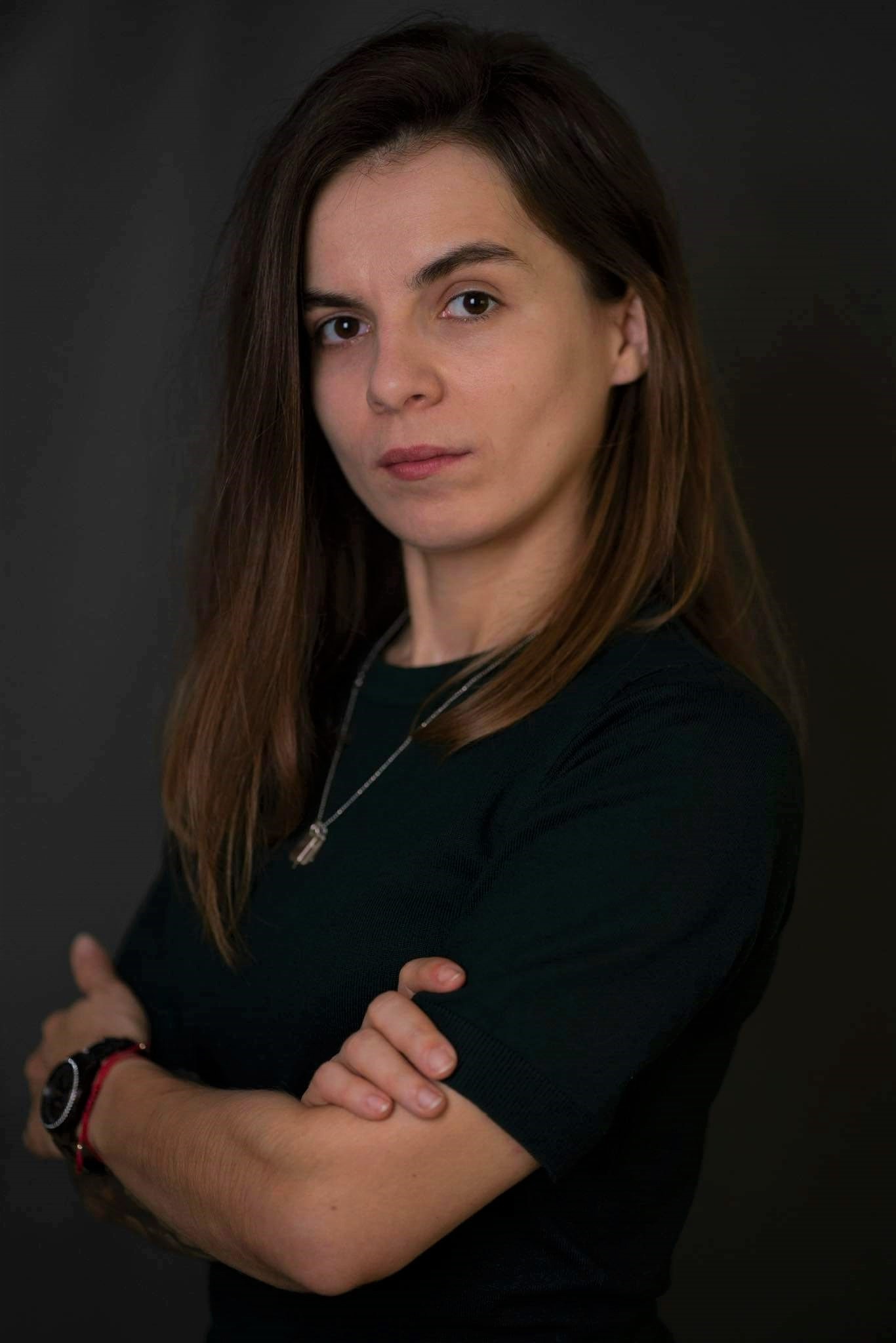 Portret kobiety z brązowymi włosami, ubrana w ciemnozielony sweter, z założonymi rękami i delikatnym naszyjnikiem na ciemnym tle.