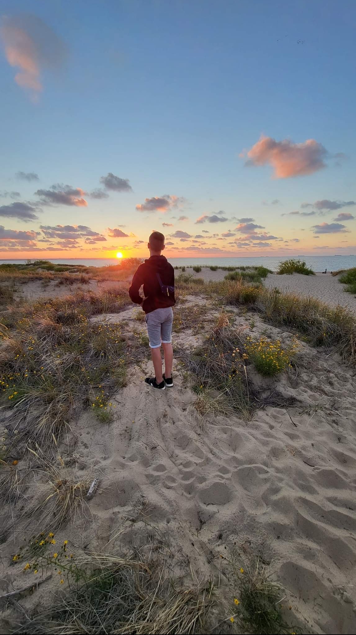 Młody mężczyzna stojący tyłem na piaszczystej wydmie, obserwujący zachód słońca nad morzem, z widocznymi chmurami zabarwionymi na różowo i żółto.