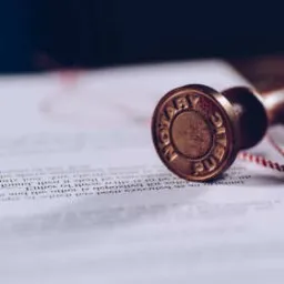 Stempel notarialny z brązową rączką i czerwonym sznurkiem leżący na dokumencie z tekstem, w tle ciemne drewno.