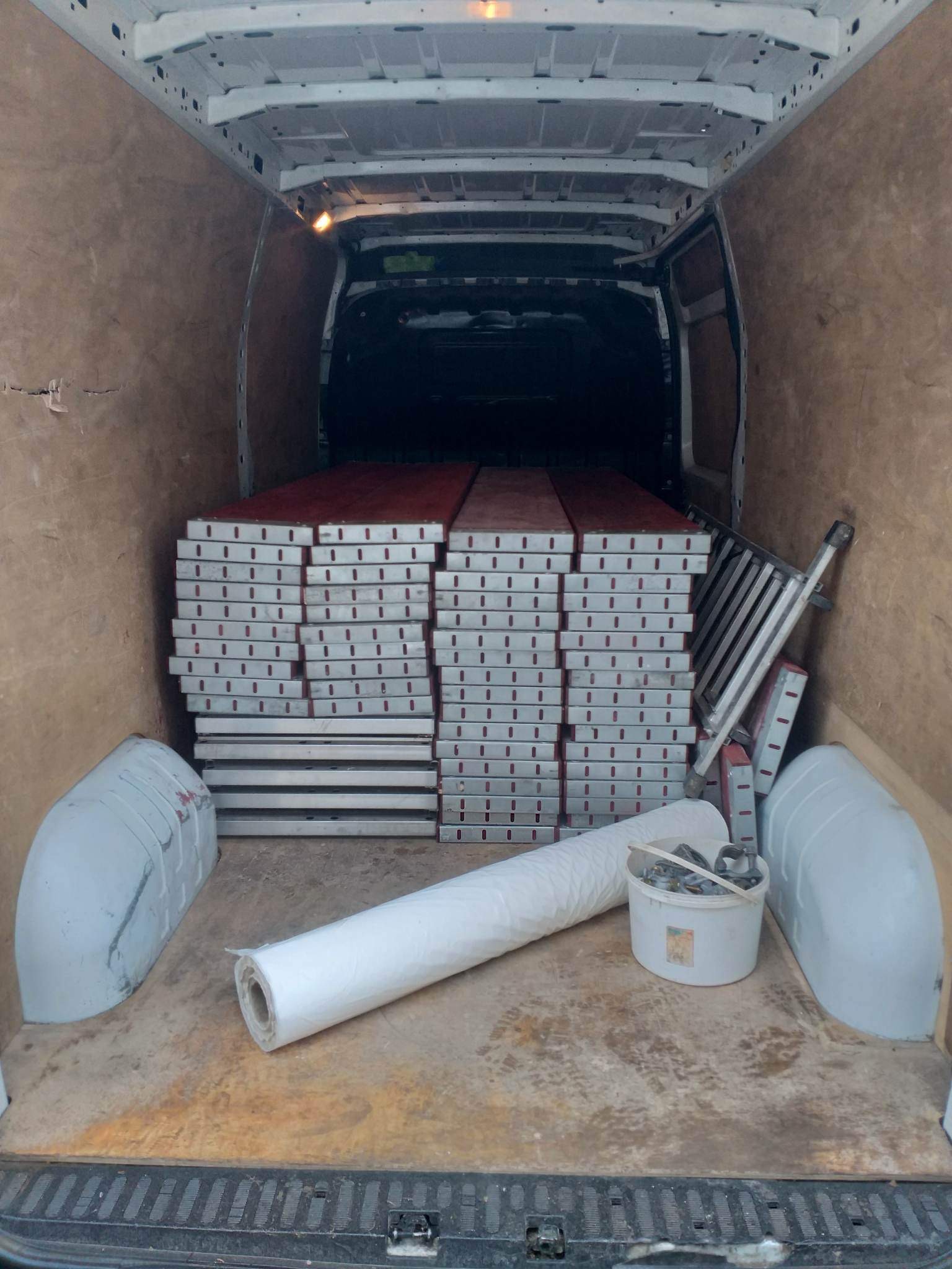 Wnętrze busa wypełnione stosem metalowych szalunków budowlanych z czerwonymi elementami, obok drabina, rolka folii i wiadro z łańcuchami, widok z tyłu pojazdu.