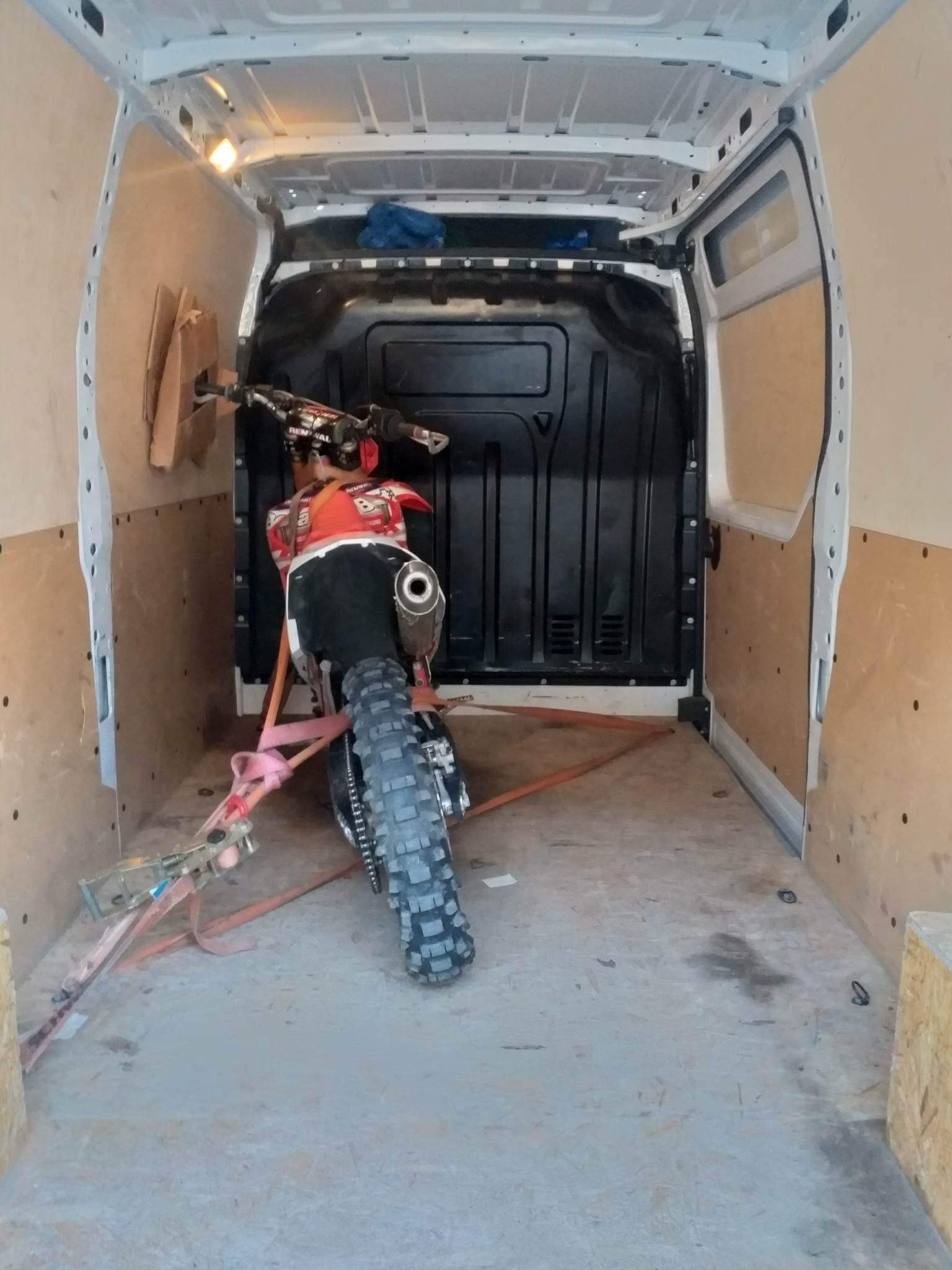 Wnętrze furgonetki z zabezpieczonym motocyklem crossowym, widoczne pasy mocujące i elementy wyposażenia pojazdu.