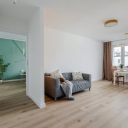 AKORD Volodymyr Rushchak - Jasne wnętrze mieszkania po remoncie, z drewnianą podłogą, szarą sofą z poduszkami i pledem, stołem z krzesłami przy oknie oraz otwartymi białymi drzwiami do pokoju z zieloną ścianą i rośliną.