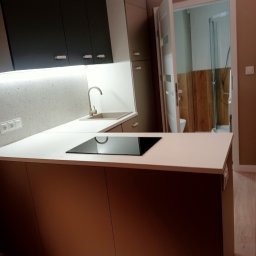 AKORD Volodymyr Rushchak - Nowoczesna kuchnia z białymi blatami, indukcyjną płytą grzewczą, jasnoszarą ścianą nad blatem, ciemnymi szafkami wiszącymi i widokiem na łazienkę z drewnianymi panelami.