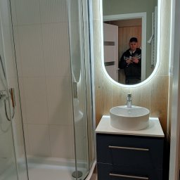 AKORD Volodymyr Rushchak - Nowoczesna łazienka po remoncie: kabina prysznicowa z białymi płytkami, umywalka nablatowa na szafce w kolorze granatowym, lustro owalne z podświetleniem LED, widoczny w odbiciu mężczyzna robiący...