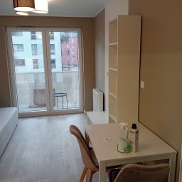 AKORD Volodymyr Rushchak - Pokój po remoncie z białymi ścianami, drewnianą podłogą, białym regałem i stołem, na którym stoją środki czystości. Widok na balkon z wyposażeniem.