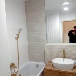 AKORD Volodymyr Rushchak - Nowoczesna łazienka po remoncie z wanną, złotą armaturą i umywalką na drewnianej szafce, widoczne akcesoria na półce w wannie i odbicie fotografa w lustrze.