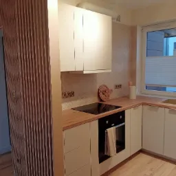 Nowoczesna kuchnia z drewnianymi akcentami: lamele na ścianie, blat, płytki w jodełkę, białe szafki i piekarnik z beżową ściereczką.