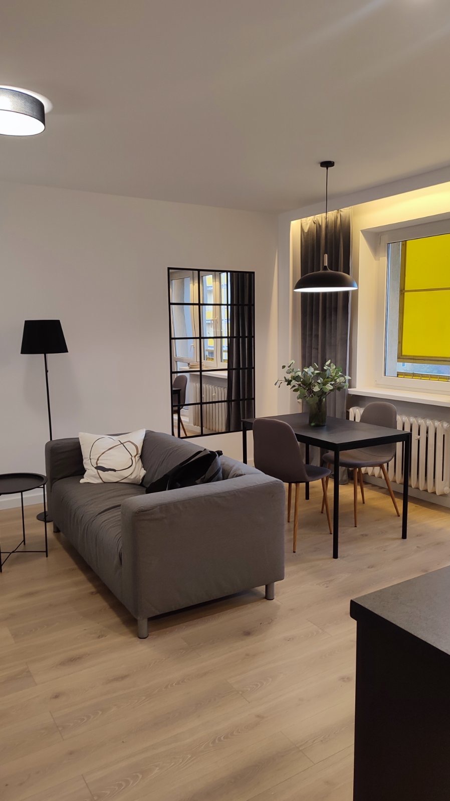 Nowoczesny salon z szarą sofą z poduszkami, czarnym lustrem w stylu industrialnym, stołem z szarymi krzesłami i designerską lampą wiszącą.