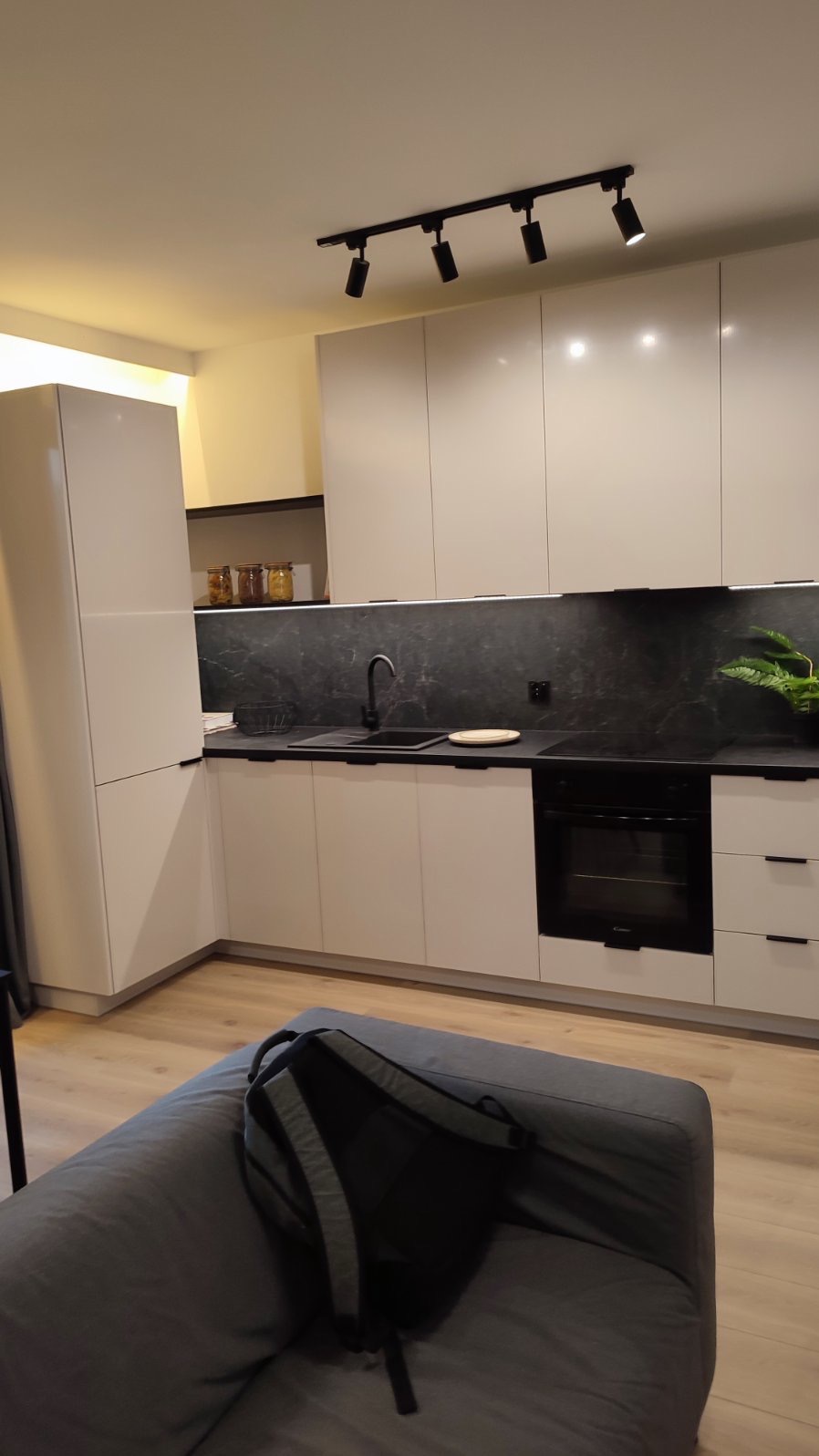 Nowoczesna kuchnia z białymi, lakierowanymi szafkami, czarnym blatem i backsplashiem o teksturze marmuru, oświetlona czarnymi reflektorami sufitowymi. Na pierwszym planie fragment szarej sofy...