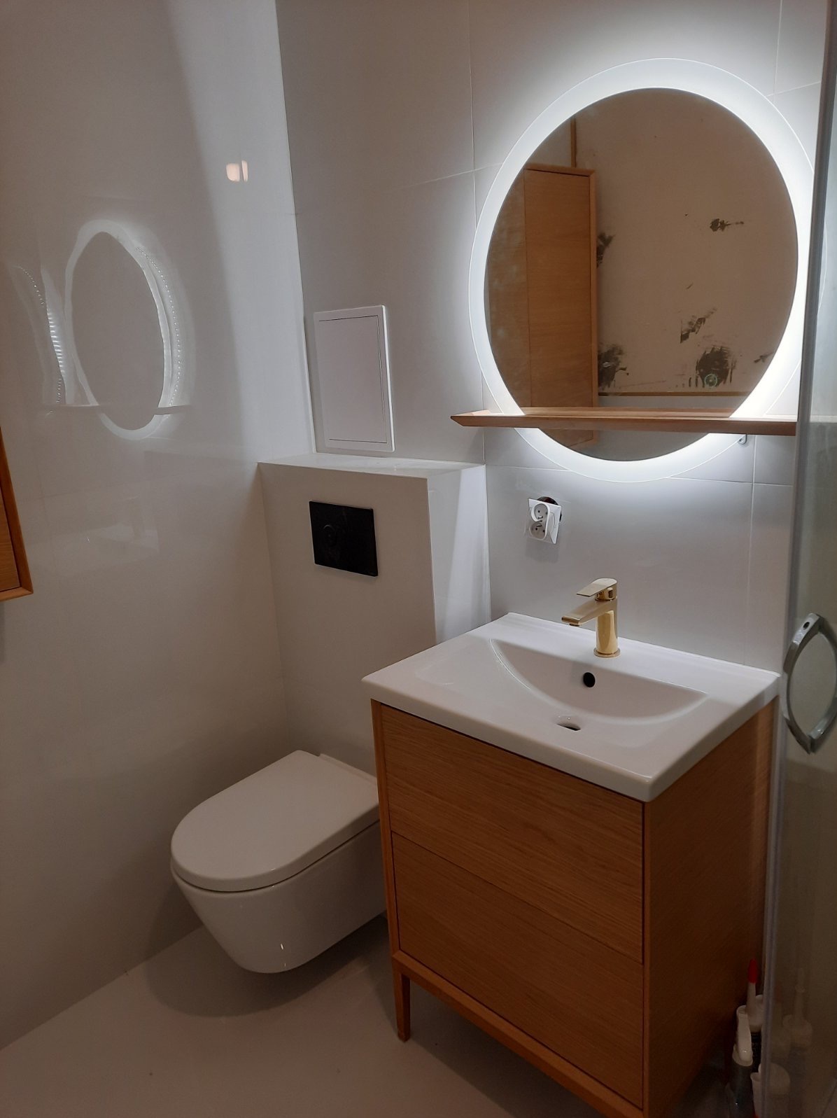 Nowoczesna łazienka z podwieszaną toaletą, umywalką na drewnianej szafce i okrągłym lustrem z podświetleniem LED, odbijającym fragment remontowanego pomieszczenia.