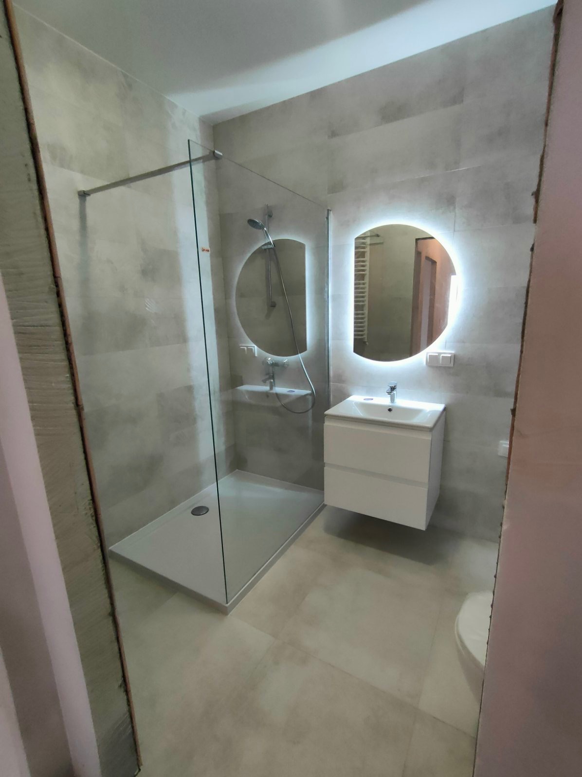 Nowoczesna łazienka z kabiną prysznicową typu walk-in, podświetlanym okrągłym lustrem i białą szafką umywalkową, wykończona w szarych płytkach imitujących beton.