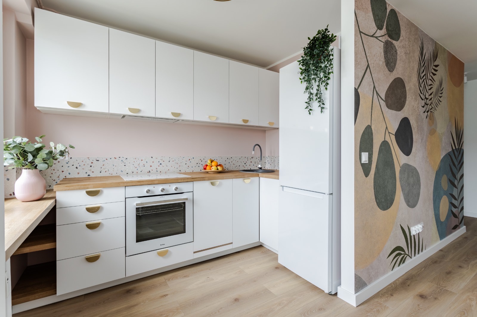 Nowoczesna kuchnia w bieli z drewnianym blatem, mozaikową ścianą nad blatem, złotymi uchwytami i dekoracyjną tapetą w roślinne wzory na ścianie bocznej.