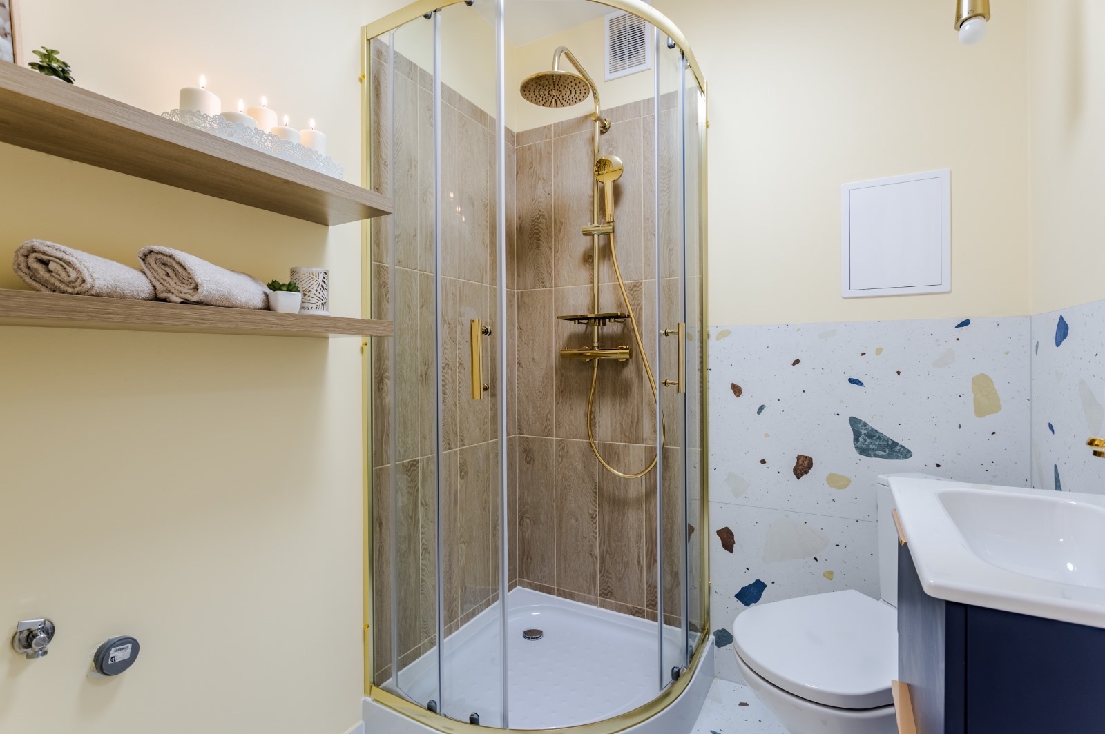 Nowoczesna łazienka z kabiną prysznicową w złotej ramie, drewnopodobnymi płytkami wewnątrz, półkami z ręcznikami i dekoracjami, oraz umywalką na tle ściany z mozaiką terrazzo.