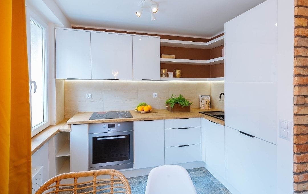 Jasna kuchnia z białymi, błyszczącymi szafkami, drewnianym blatem, płytą indukcyjną i piekarnikiem, uzupełniona ceglaną ścianą i żółtą zasłoną w oknie.