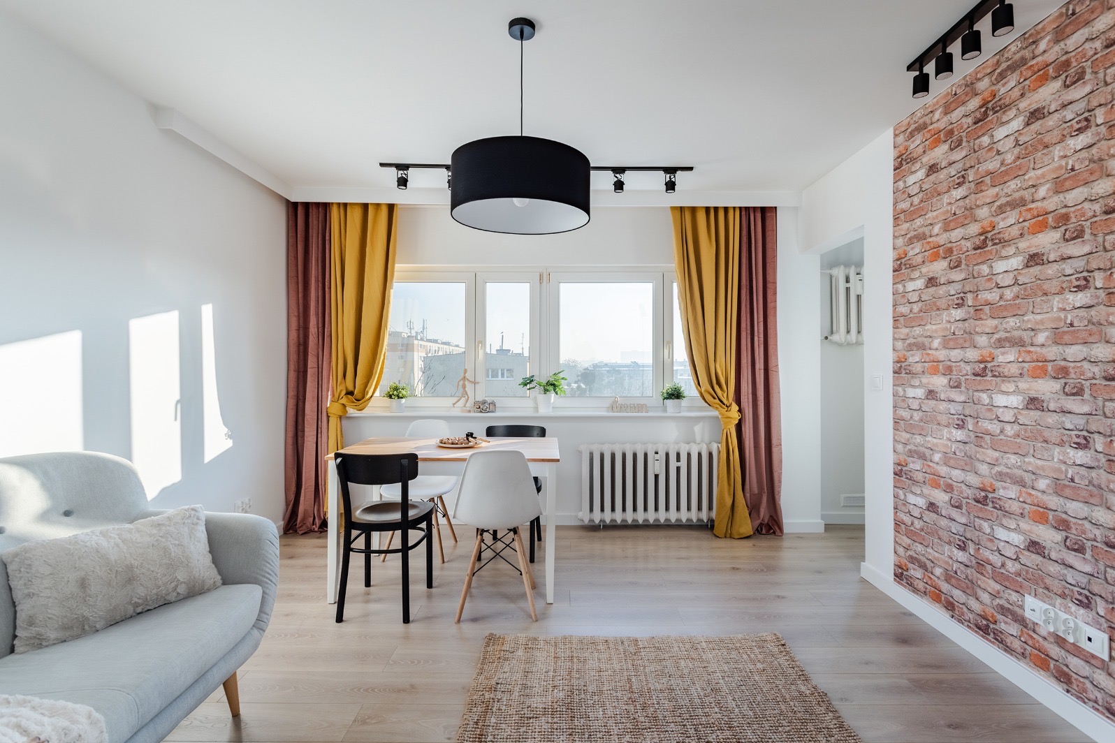 Jasne wnętrze z ceglaną ścianą, drewnianym stołem, krzesłami w stylu skandynawskim i zasłonami w odcieniach złota i bordo, oświetlone lampą sufitową z abażurem