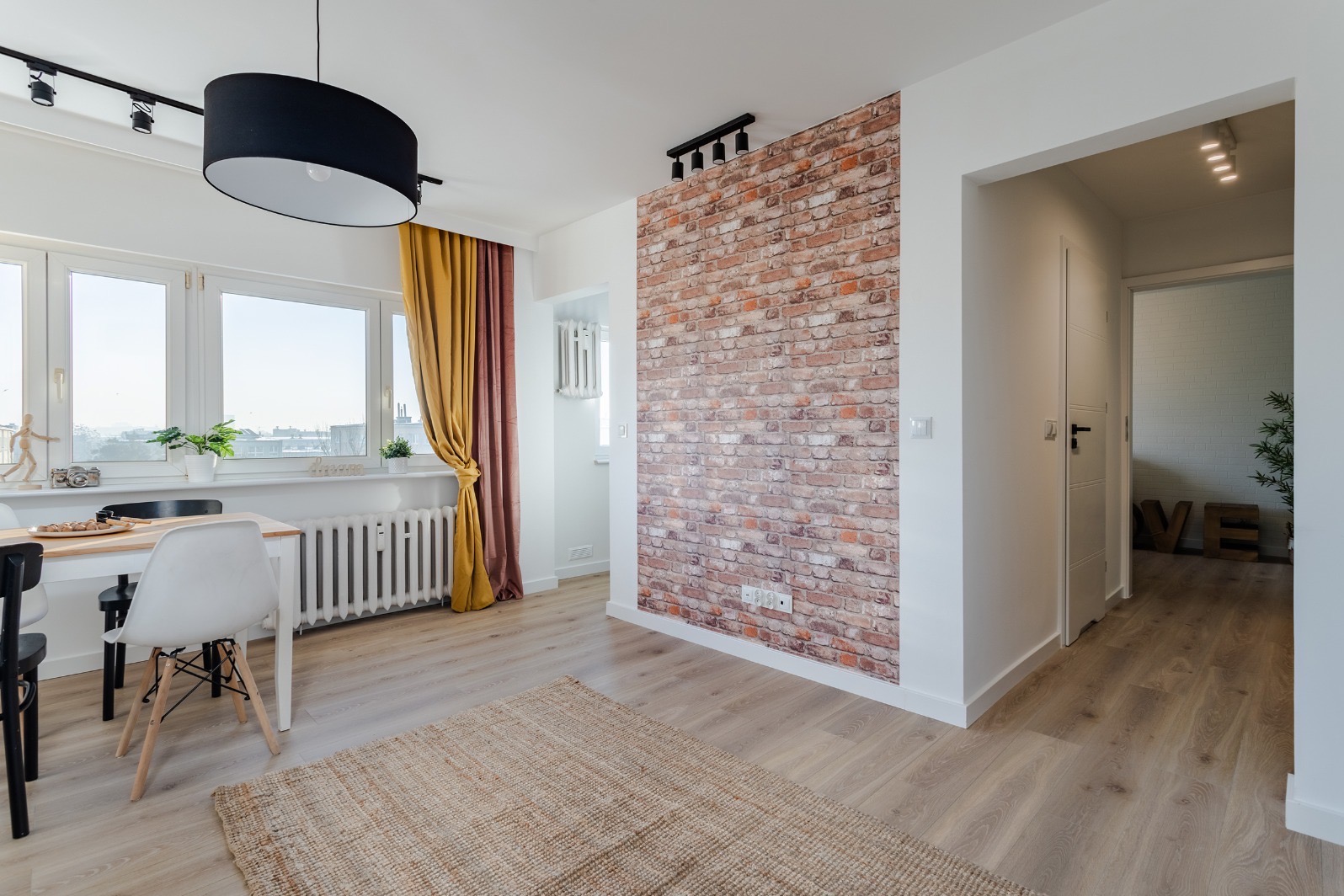 Jasne wnętrze mieszkania z drewnianą podłogą, ceglaną ścianą akcentującą, stołem jadalnym pod oknem i złotymi zasłonami.