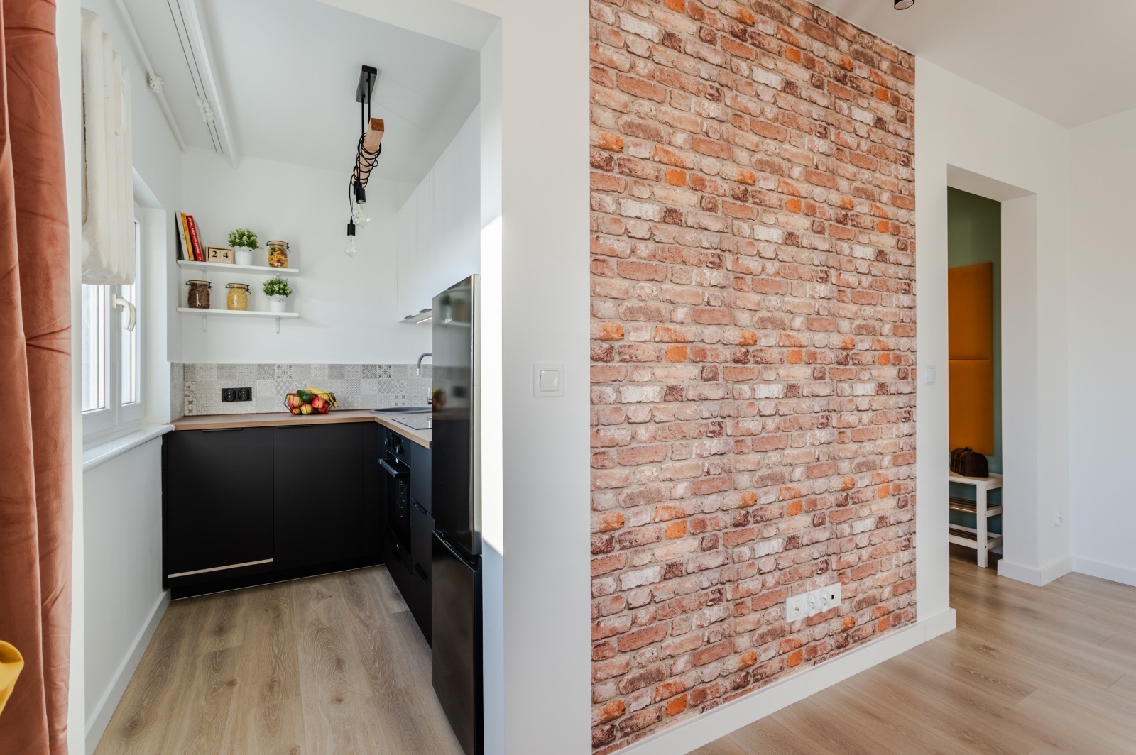Nowoczesna kuchnia z czarnymi szafkami, drewnianym blatem i ceglaną ścianą dekoracyjną, widok z przedpokoju
