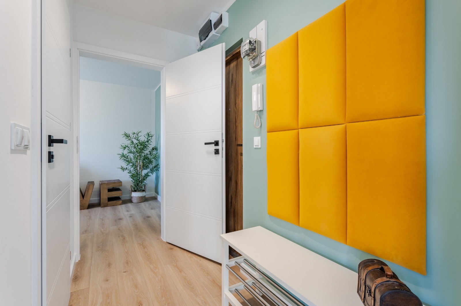 Jasny przedpokój z otwartymi białymi drzwiami, drewnianą podłogą, żółtym panelem ściennym, białą szafką na buty i dekoracyjną skrzynią