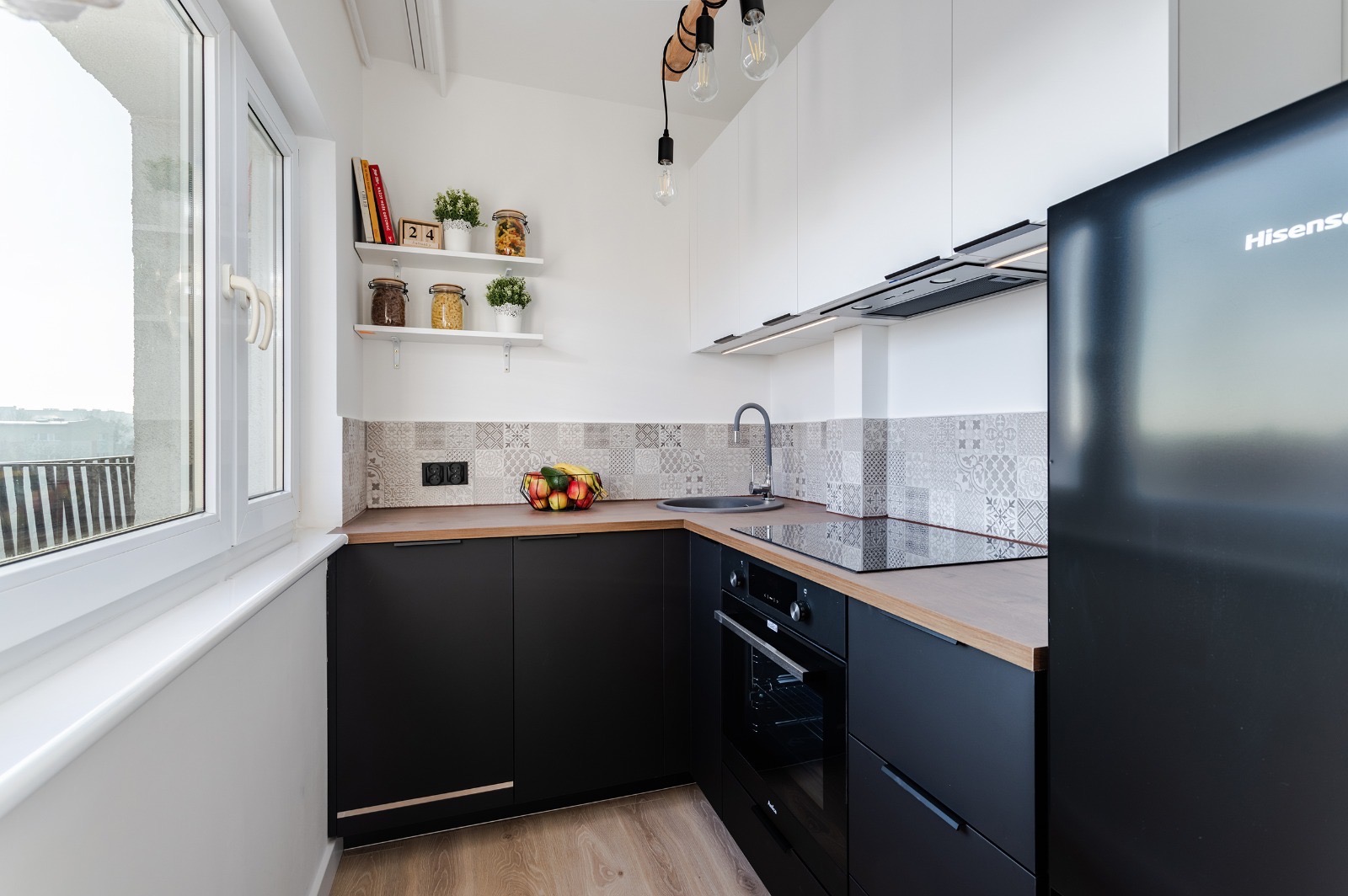 Nowoczesna kuchnia w czarno-białej kolorystyce z drewnianymi akcentami, widok na okno z parapetem i balkon, dekoracyjne płytki nad blatem, oświetlenie wiszące z żarówkami Edisona.