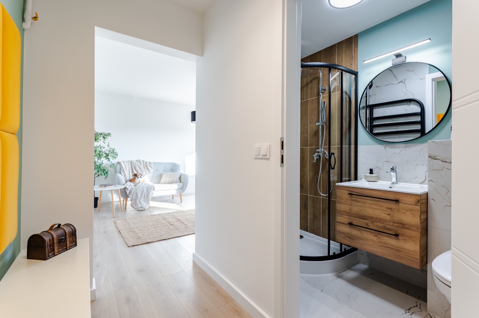 Nowoczesna łazienka z kabiną prysznicową z czarnymi profilami, drewnianą szafką pod umywalką i okrągłym lustrem, widok z korytarza z elementami dekoracyjnymi w stylu retro.