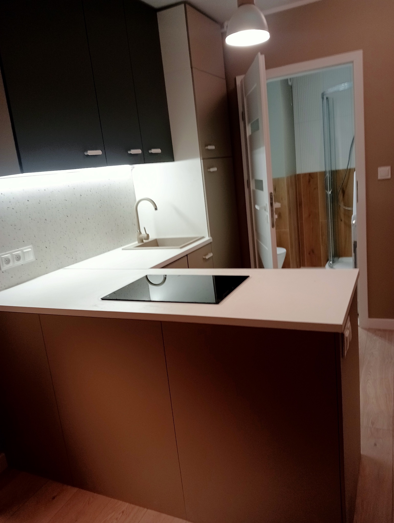 Nowoczesna kuchnia z białymi blatami, indukcyjną płytą grzewczą, jasnoszarą ścianą nad blatem, ciemnymi szafkami wiszącymi i widokiem na łazienkę z drewnianymi panelami.