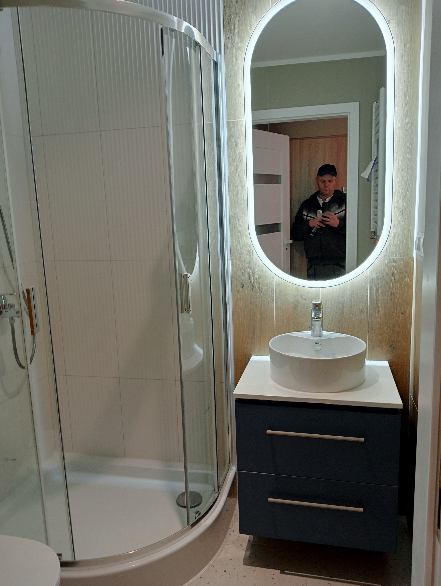 Nowoczesna łazienka po remoncie: kabina prysznicowa z białymi płytkami, umywalka nablatowa na szafce w kolorze granatowym, lustro owalne z podświetleniem LED, widoczny w odbiciu mężczyzna robiący...