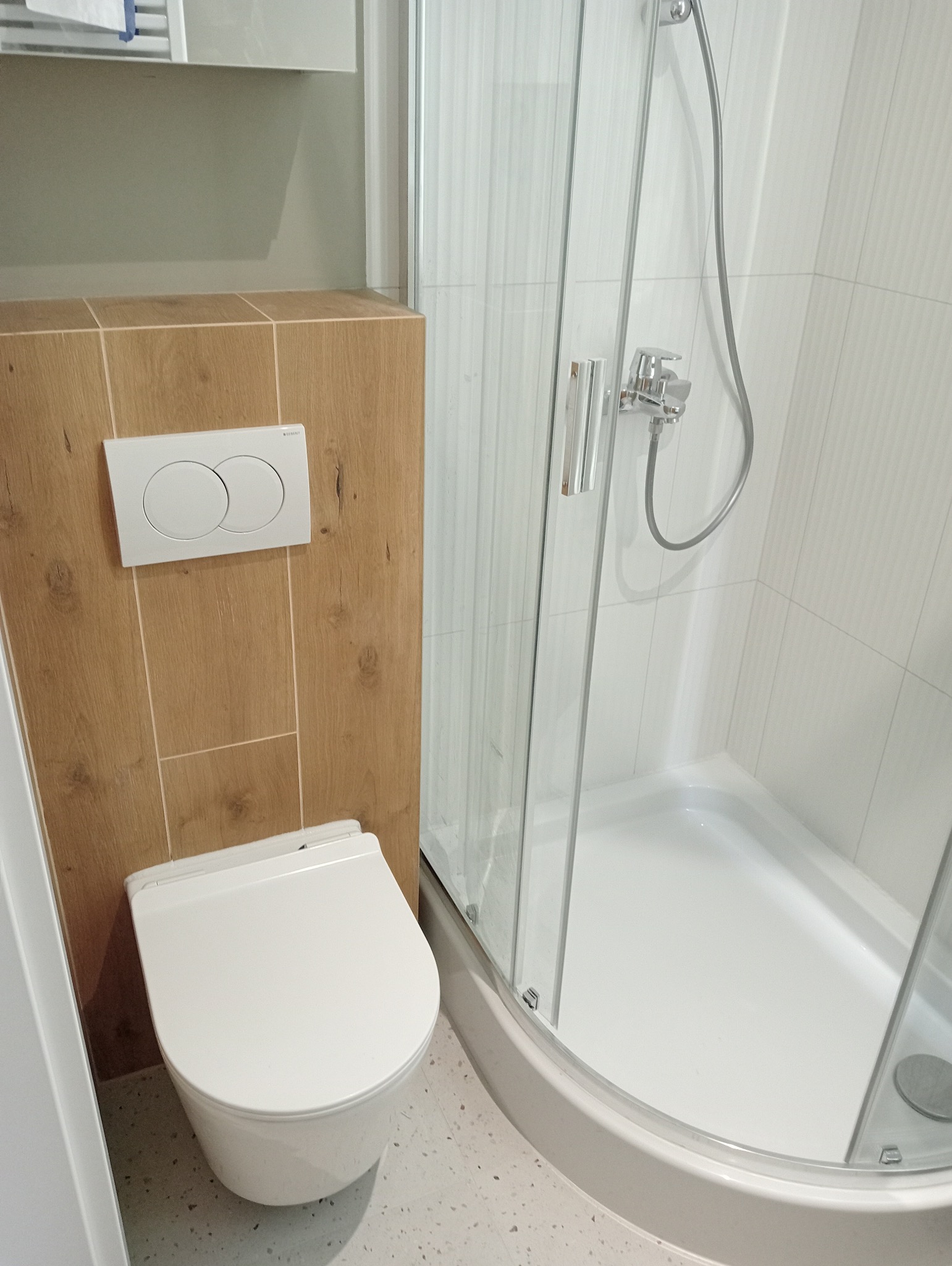 Nowoczesna łazienka z podwieszaną toaletą i kabiną prysznicową z białymi płytkami imitującymi drewno na obudowie toalety.