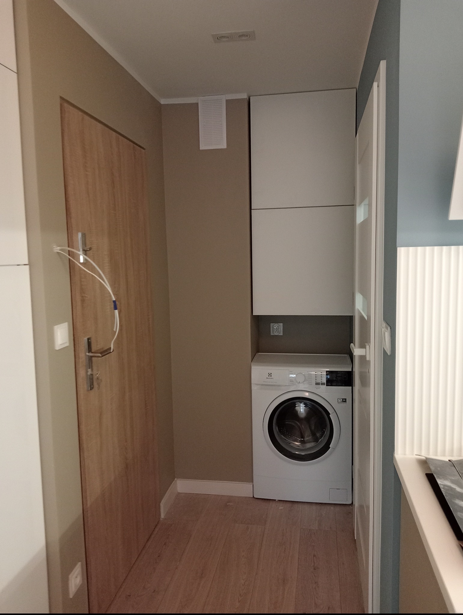 Niewielka łazienka po remoncie: pralka Electrolux w zabudowie z białymi szafkami, drewniana podłoga i drzwi z metalowymi klamkami.