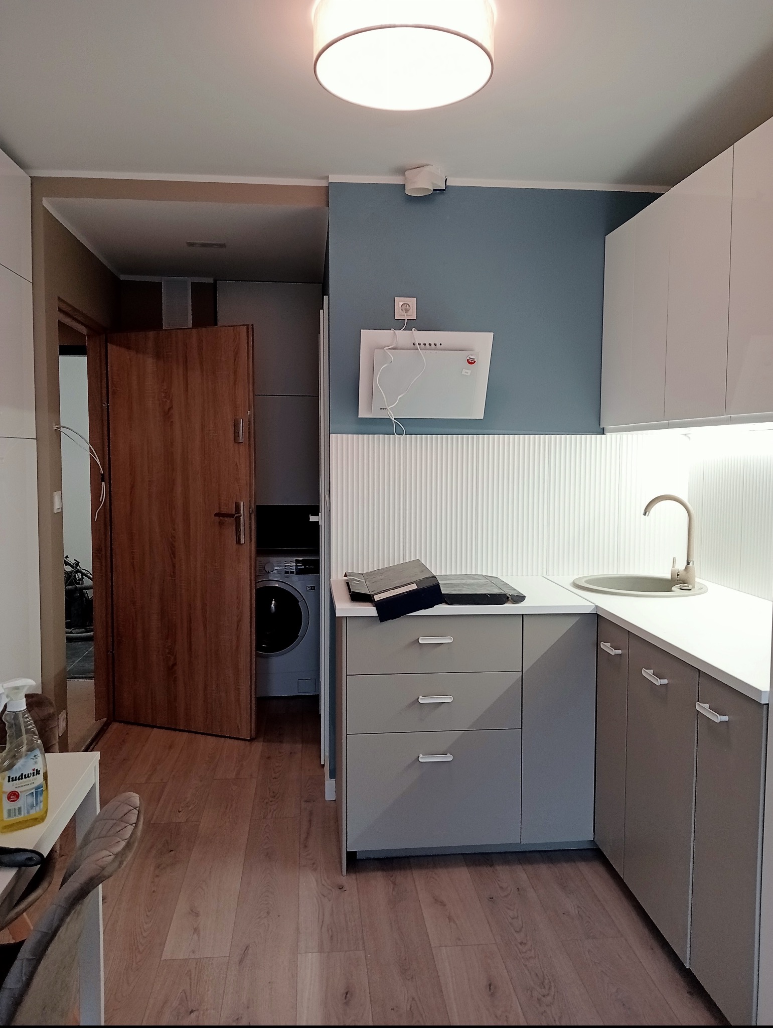 Nowoczesna kuchnia w odcieniach szarości i błękitu z drewnianą podłogą i drzwiami, widoczna pralka w tle, na blacie leżą narzędzia.