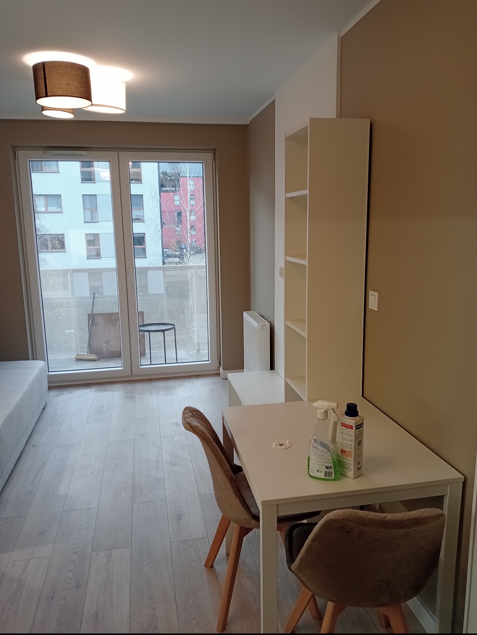 Pokój po remoncie z białymi ścianami, drewnianą podłogą, białym regałem i stołem, na którym stoją środki czystości. Widok na balkon z wyposażeniem.