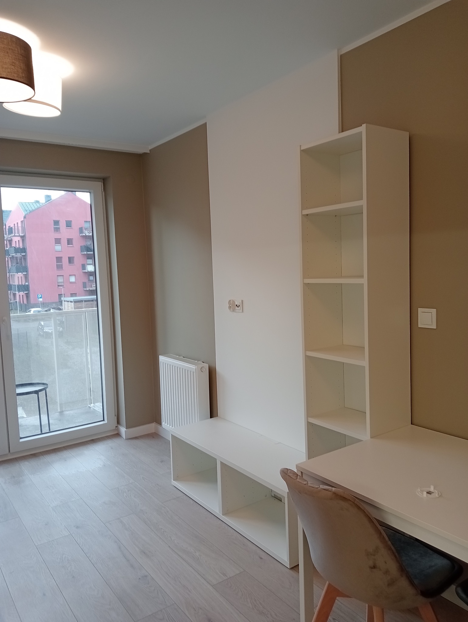 Jasne wnętrze pokoju z białymi meblami: regał, biurko i szafka pod telewizor, z widokiem na balkon i różowy budynek za oknem.