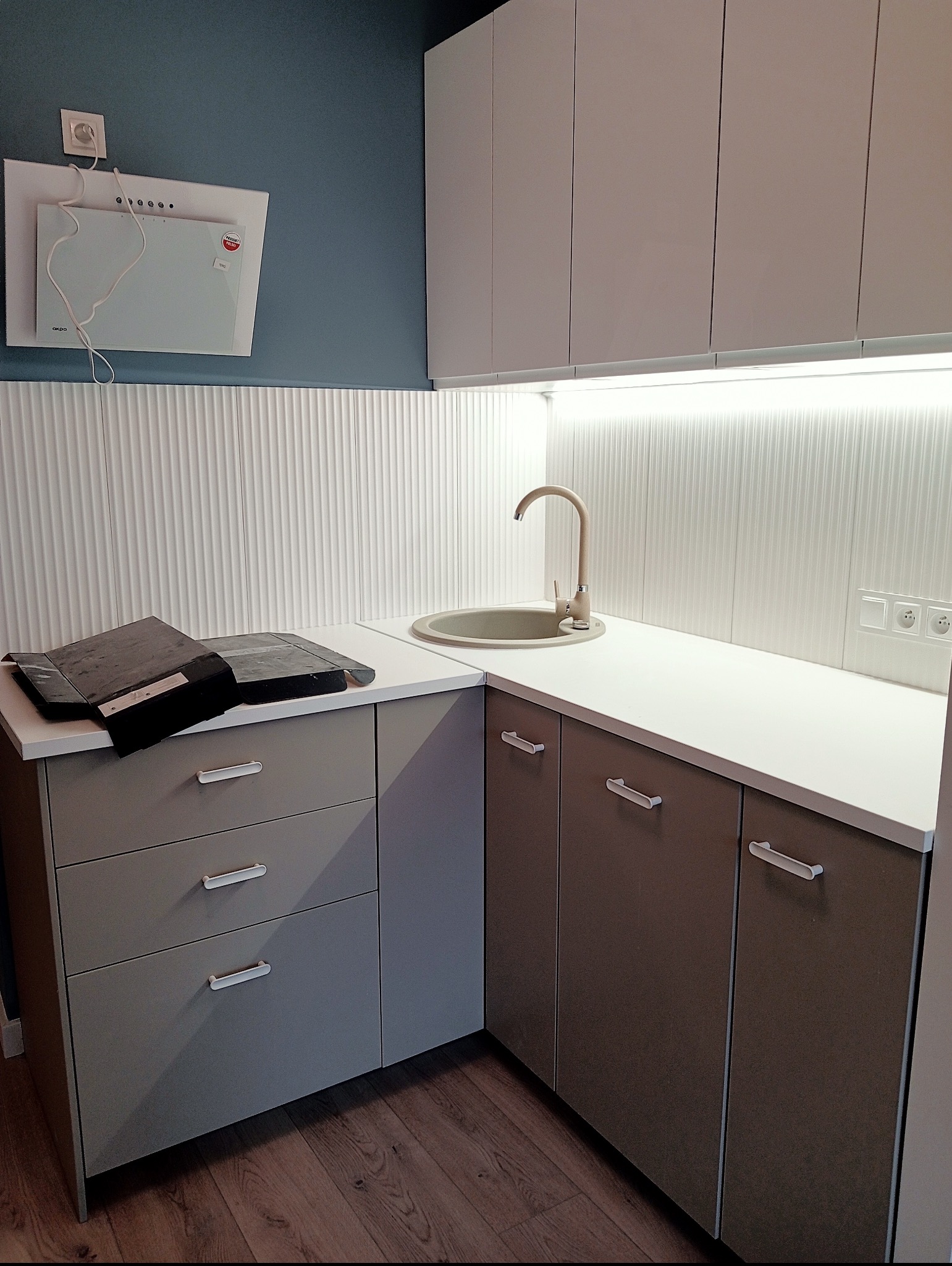 Nowoczesna kuchnia z beżowym zlewem i baterią, białym blatem oraz szarymi szafkami z podłużnymi uchwytami, na blacie leżą zabezpieczenia przed zabrudzeniem w trakcie remontu.