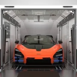 Pomarańczowy sportowy samochód z czarną maską umieszczony wewnątrz białej, zamkniętej przyczepy z rampą najazdową. Widoczny przód pojazdu i elementy konstrukcyjne przyczepy.