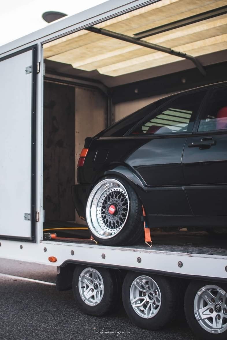 Czarny samochód osobowy, z widocznymi felgami BBS, zabezpieczony pomarańczowymi pasami, transportowany wewnątrz białej przyczepy z trzema osiami.