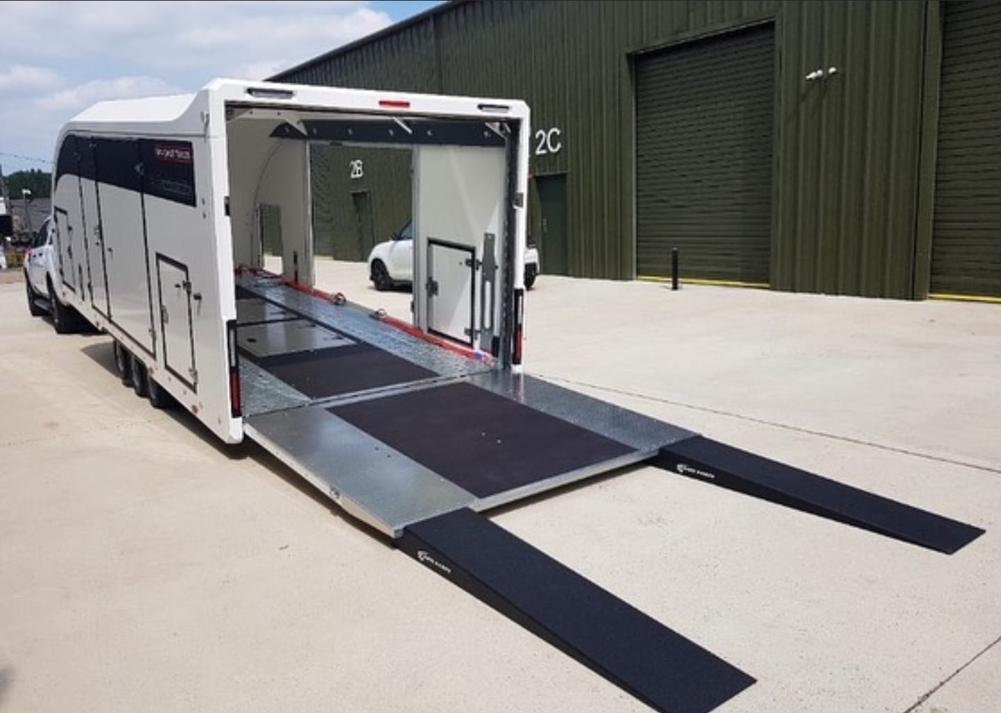 Otwarta laweta samochodowa z wysuniętymi rampami najazdowymi, widoczny samochód wewnątrz oraz budynek magazynowy w tle.