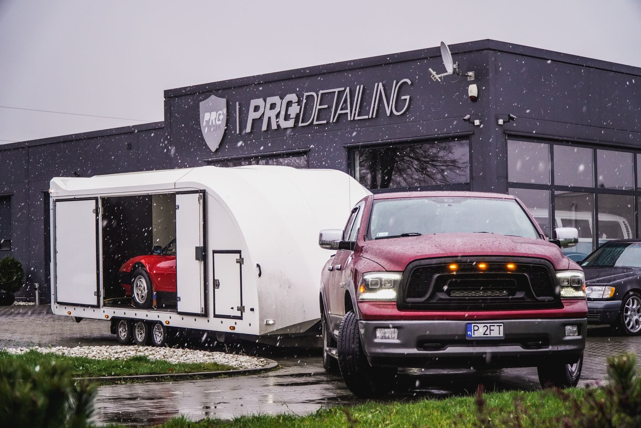 Czerwony samochód sportowy w białej zabudowanej przyczepie transportowej, ciągniętej przez bordowego Dodge Ram, na tle szarego budynku z logo 'PRG Detailing' podczas opadów śniegu.