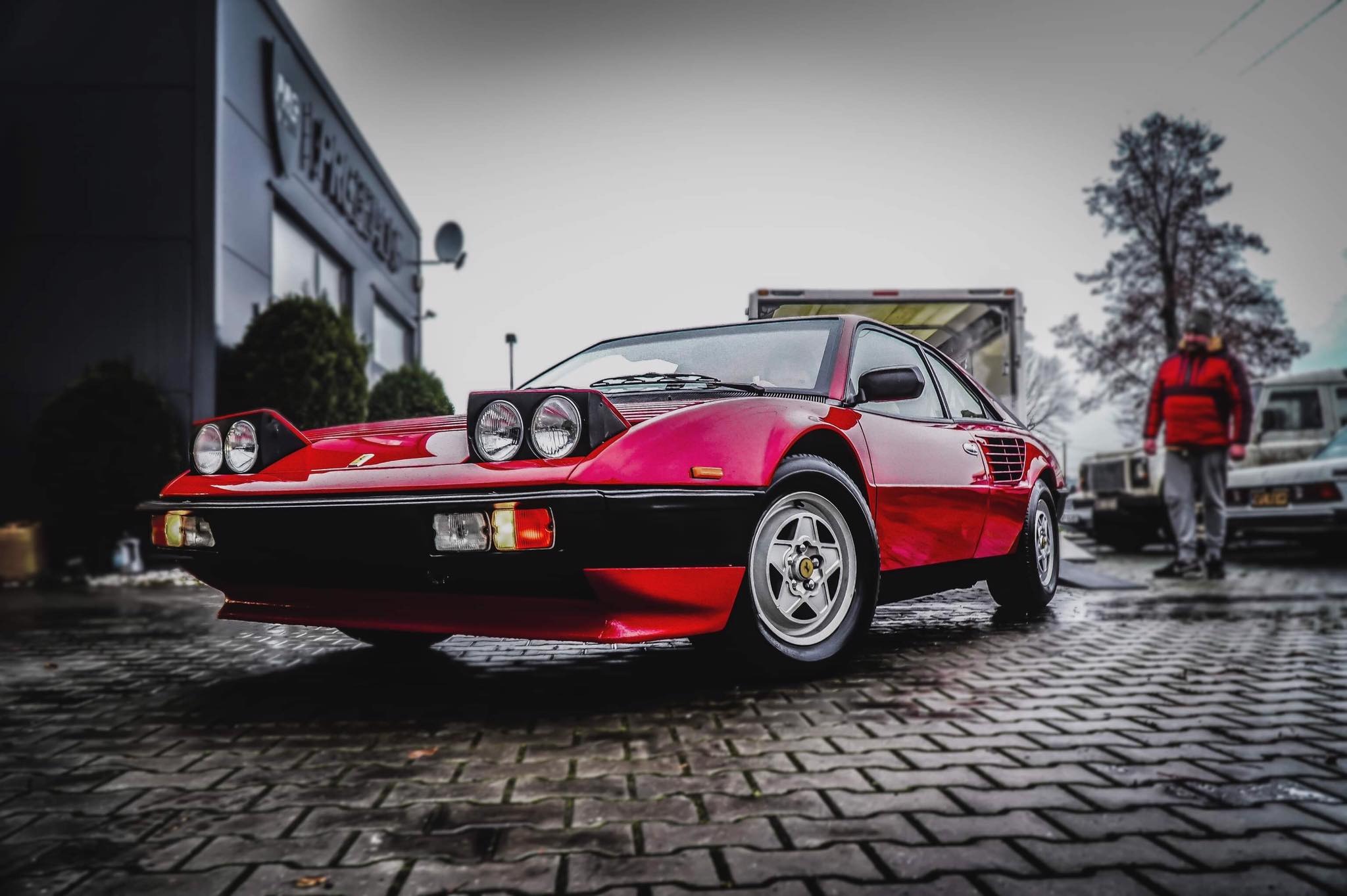 Czerwone Ferrari 308 GT4 z dodatkowymi reflektorami na masce, zaparkowane na mokrej kostce brukowej przed nowoczesnym budynkiem, w tle postać w czerwonej kurtce i przyczepa.