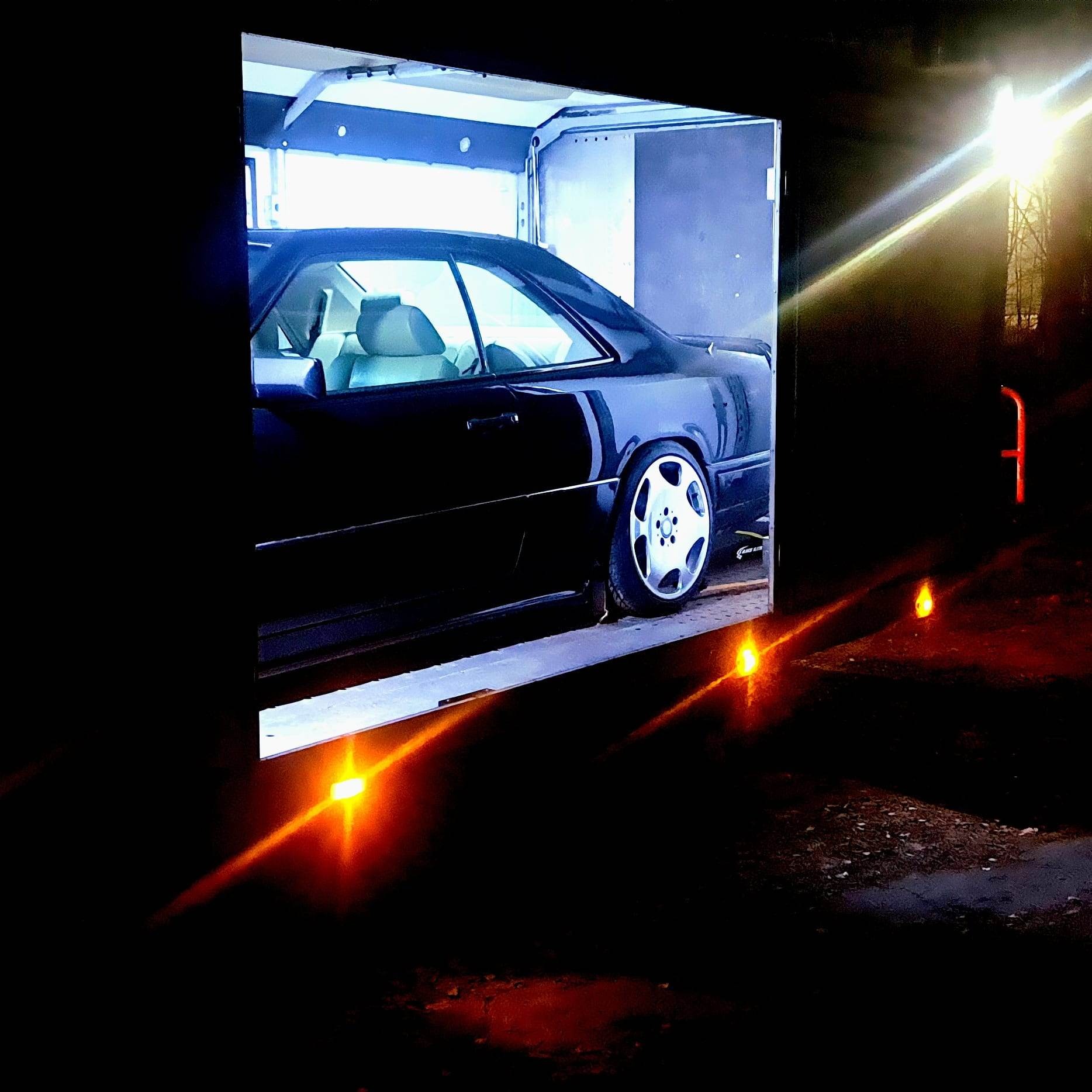 Czarny samochód coupe Mercedes-Benz W124 na platformie lawety w nocy, oświetlony pomarańczowymi światłami obrysowymi.