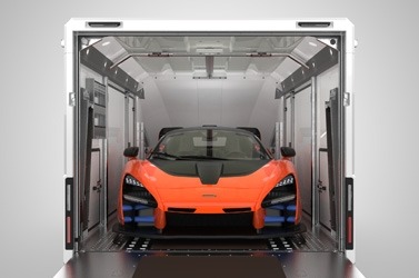 Pomarańczowy sportowy samochód z czarną maską umieszczony wewnątrz białej, zamkniętej przyczepy z rampą najazdową. Widoczny przód pojazdu i elementy konstrukcyjne przyczepy.