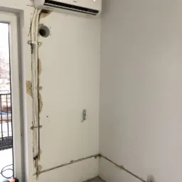 Nowoczesny klimatyzator ścienny zamontowany nad oknem, widoczne kable i rury prowadzone po ścianie w trakcie montażu, ślady po kuciu w ścianie.