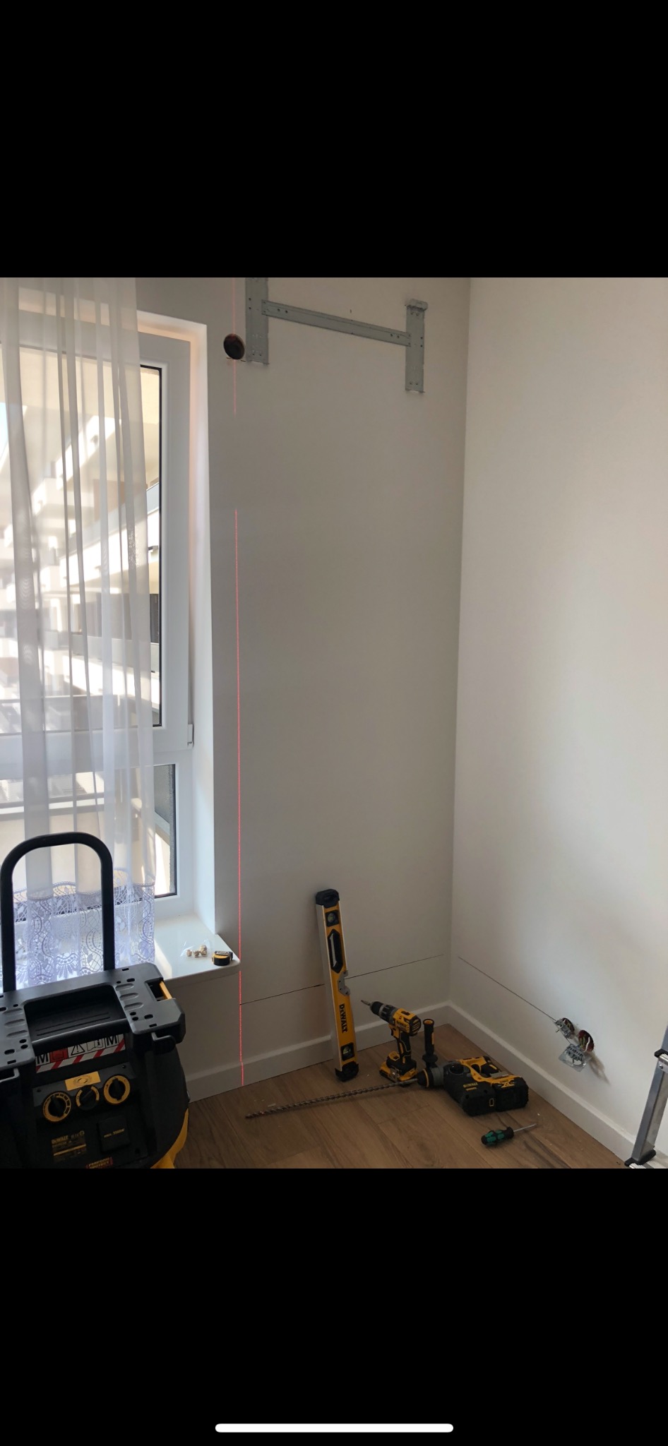 Montaż klimatyzacji: wsporniki zamocowane na ścianie, poziomica laserowa wyznacza pion, narzędzia DeWalt na podłodze, widoczne przewody elektryczne.