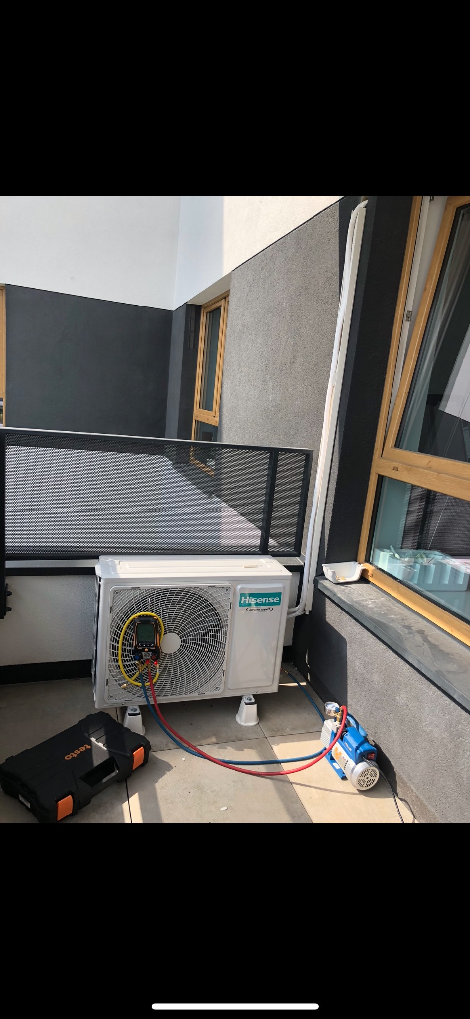 Biała jednostka zewnętrzna klimatyzacji Hisense zamontowana na balkonie, podłączona do pompy próżniowej i manometru cyfrowego Testo, widoczna czarna skrzynka narzędziowa.
