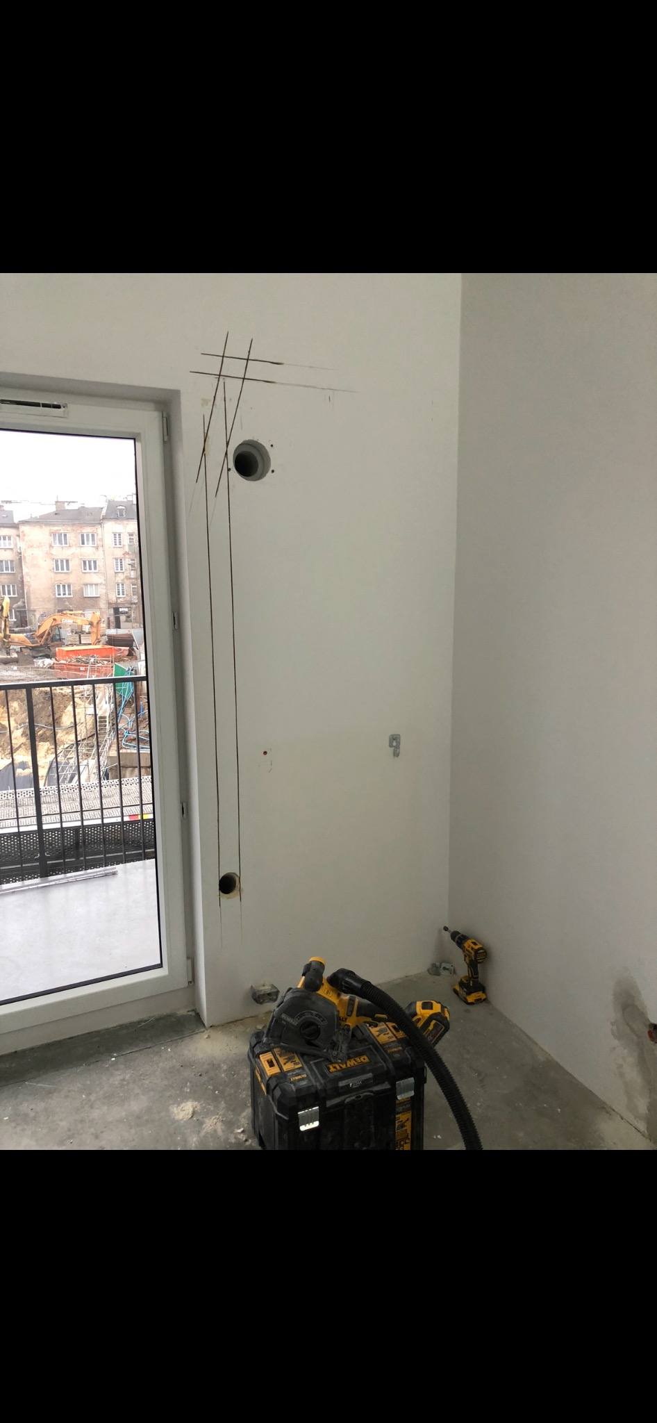 Przygotowanie do instalacji: Wywiercone otwory w ścianie, zaznaczone linie, skrzynka narzędziowa DeWalt, wiertarka i odkurzacz przemysłowy w surowym wnętrzu z widokiem na plac budowy.