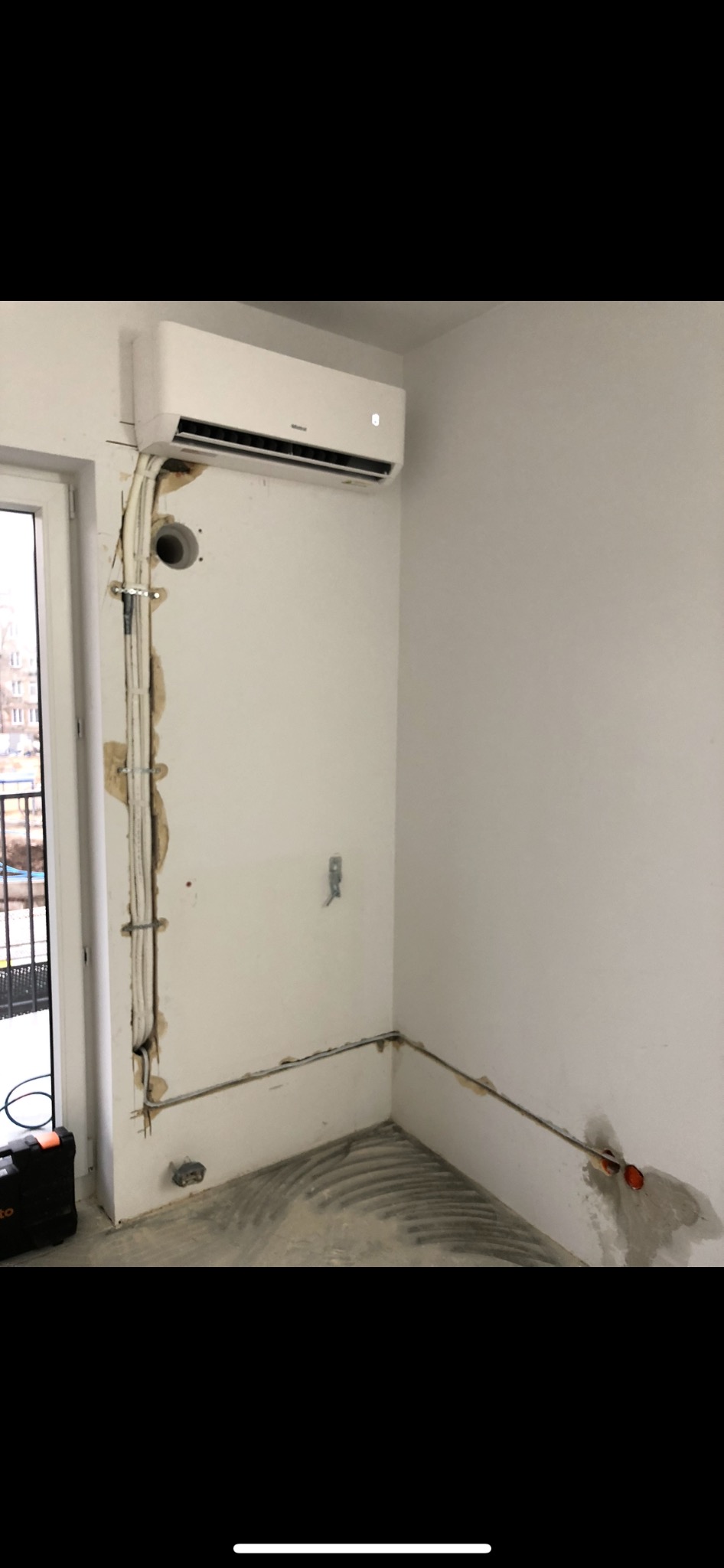 Nowoczesny klimatyzator ścienny zamontowany nad oknem, widoczne kable i rury prowadzone po ścianie w trakcie montażu, ślady po kuciu w ścianie.