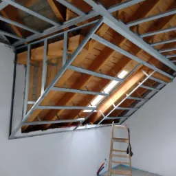 Konstrukcja metalowa podwieszanego sufitu z widoczną izolacją z desek na poddaszu, drabina oparta o ścianę.