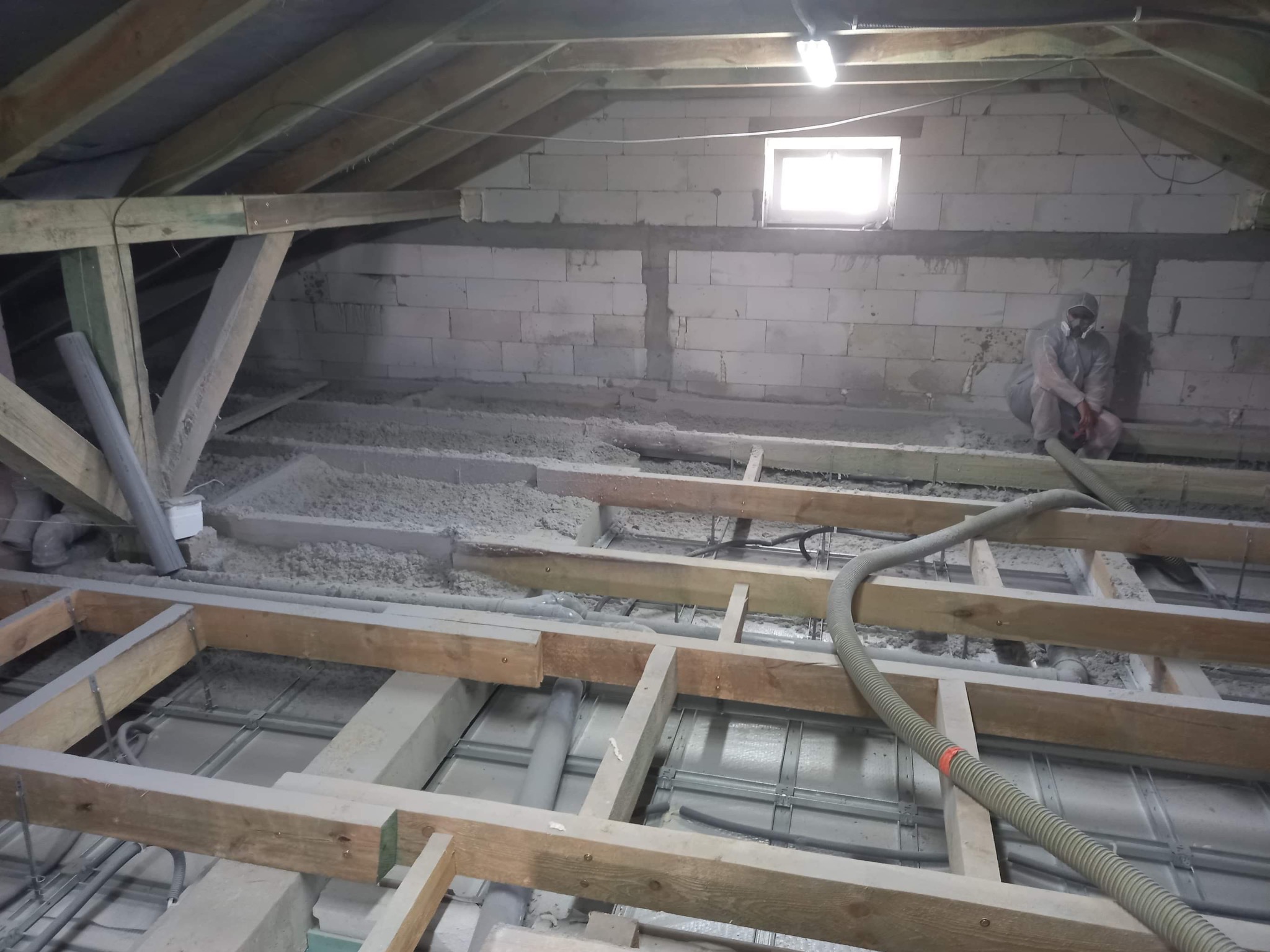 Poddasze w trakcie izolacji: widoczne drewniane legary, szara izolacja między nimi oraz pracownik w kombinezonie ochronnym siedzący na ścianie z bloczków betonowych, obok okno.