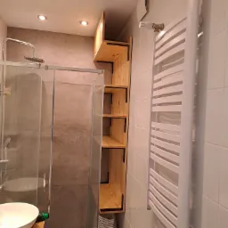 Wnętrze łazienki z drewnianą p&oacute;łką wiszącą, kabiną prysznicową i grzejnikiem drabinkowym na białej ścianie z płytek.