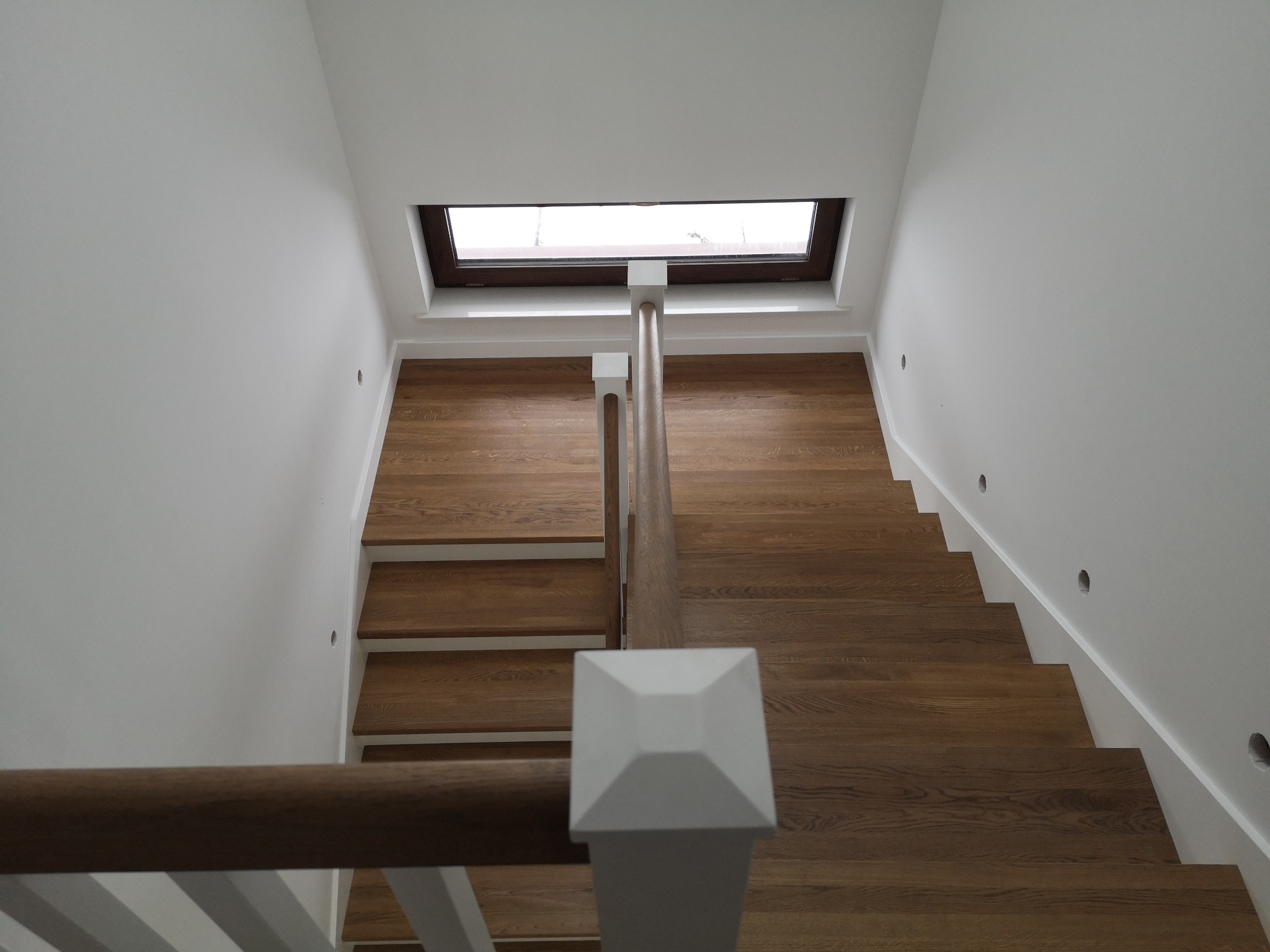 Widok z góry na nowoczesne, dębowe schody z drewnianą balustradą i białymi słupkami, oświetlone przez okno dachowe w minimalistycznym, białym wnętrzu.