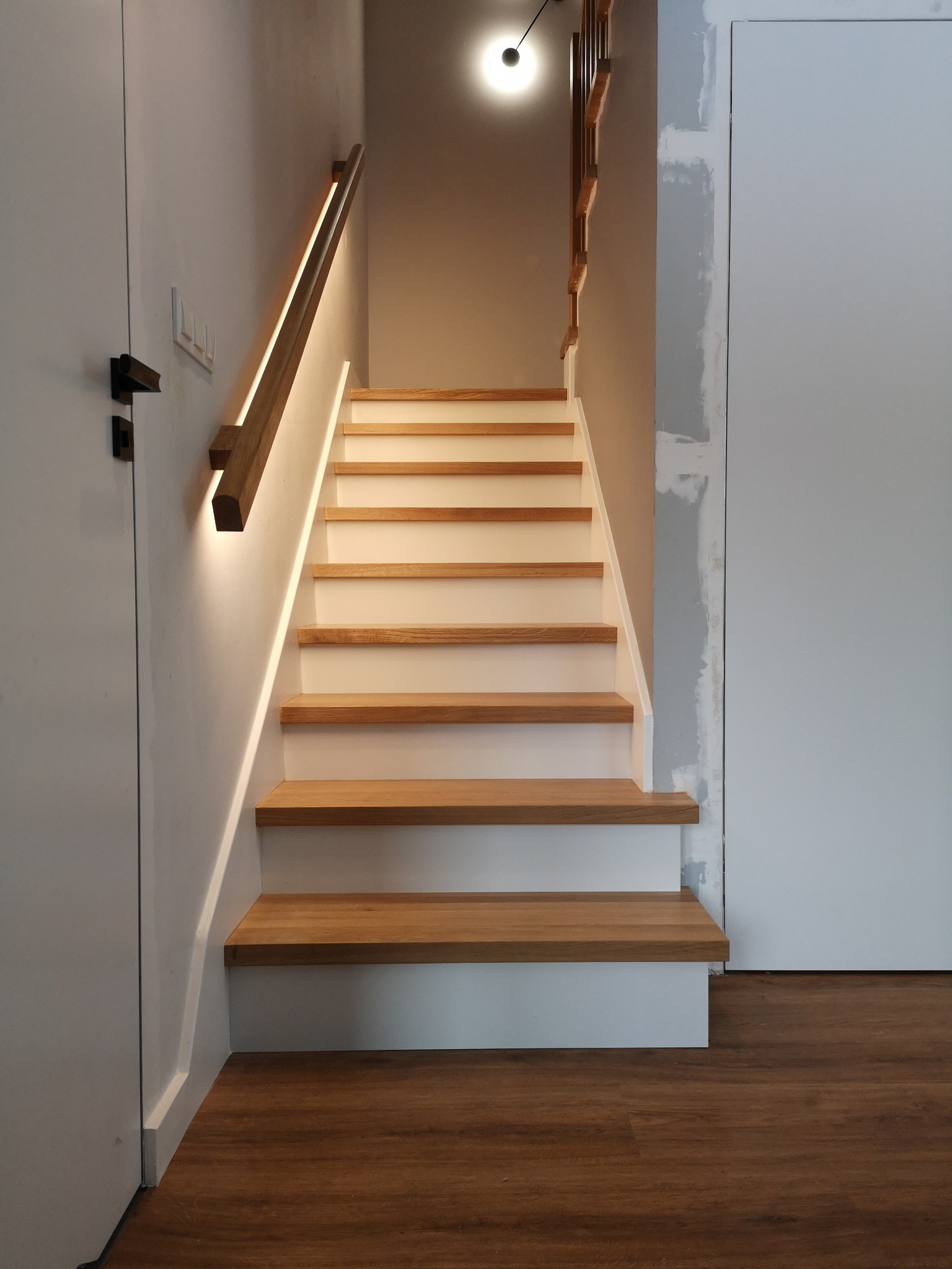 Bieg schodów z drewnianymi stopniami i białymi podstopnicami, oświetlony listwą LED wzdłuż poręczy, widok z dołu na górę.